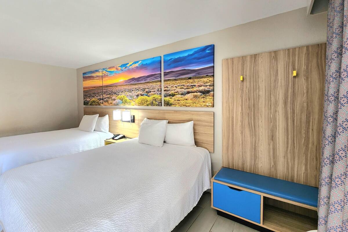 A hotel room with two beds, a wooden wardrobe, a blue bench, and a large landscape painting above the beds.
