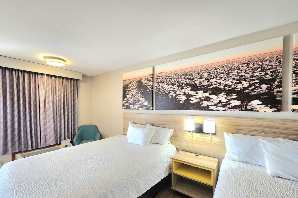 A hotel room with two beds, a headboard with wall sconces, a large floral artwork above the beds, and a window with curtains.