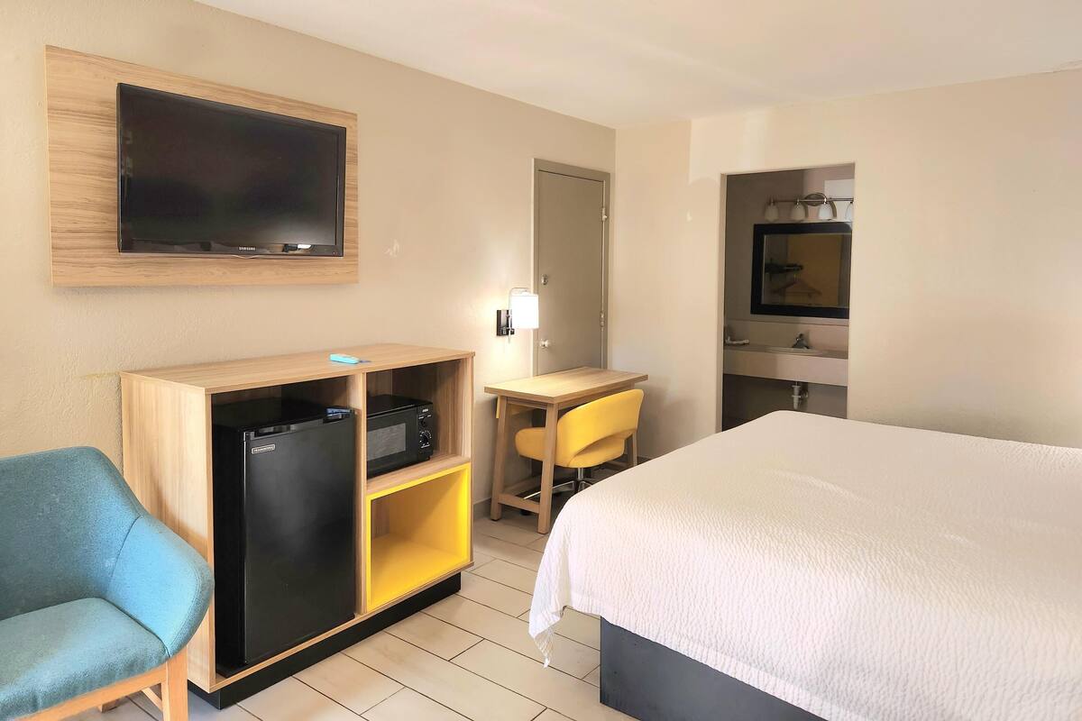 A hotel room with a bed, a desk with a chair, a television mounted on the wall, a refrigerator, and a mirror.