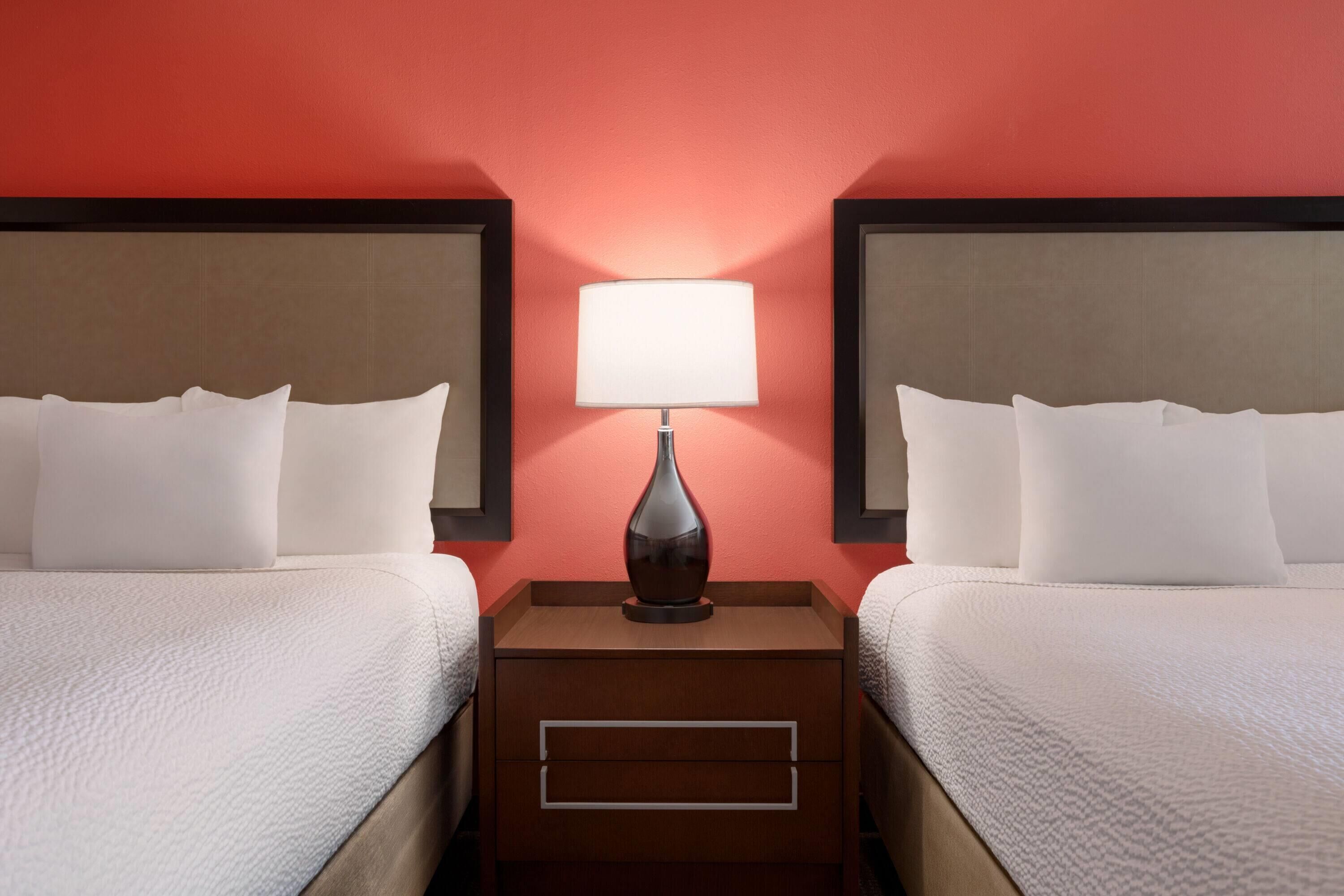 A hotel room with two beds, a nightstand with a lamp, and a red wall.