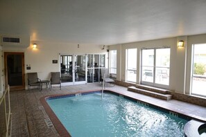 An indoor swimming pool with clear water, a diving board, and a seating area with chairs.