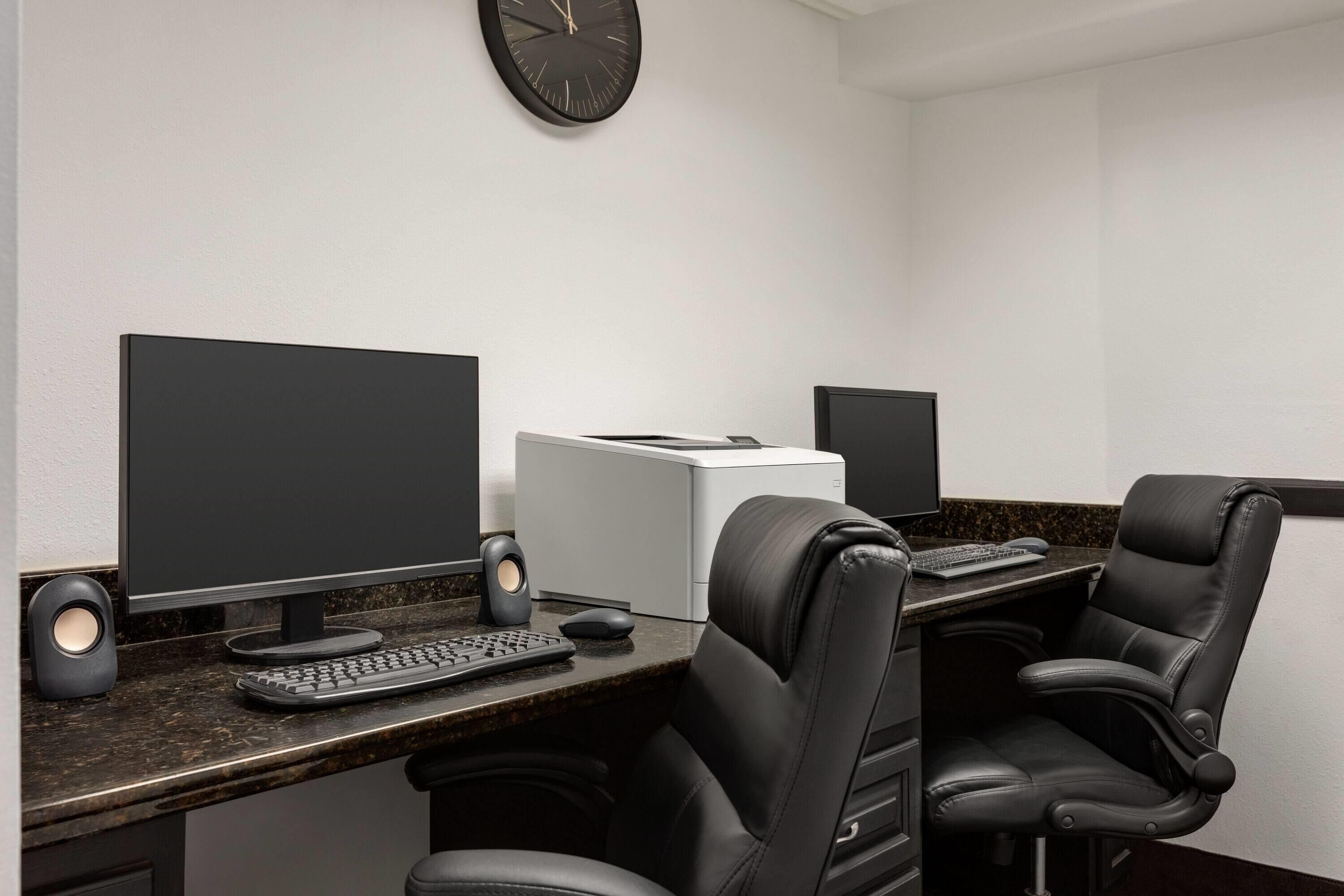 Un espacio de oficina moderno con dos escritorios, cada uno equipado con un monitor de computadora, teclado y altavoces. Hay dos sillas de cuero negro, un reloj en la pared e impresora blanca sobre los escritorios.