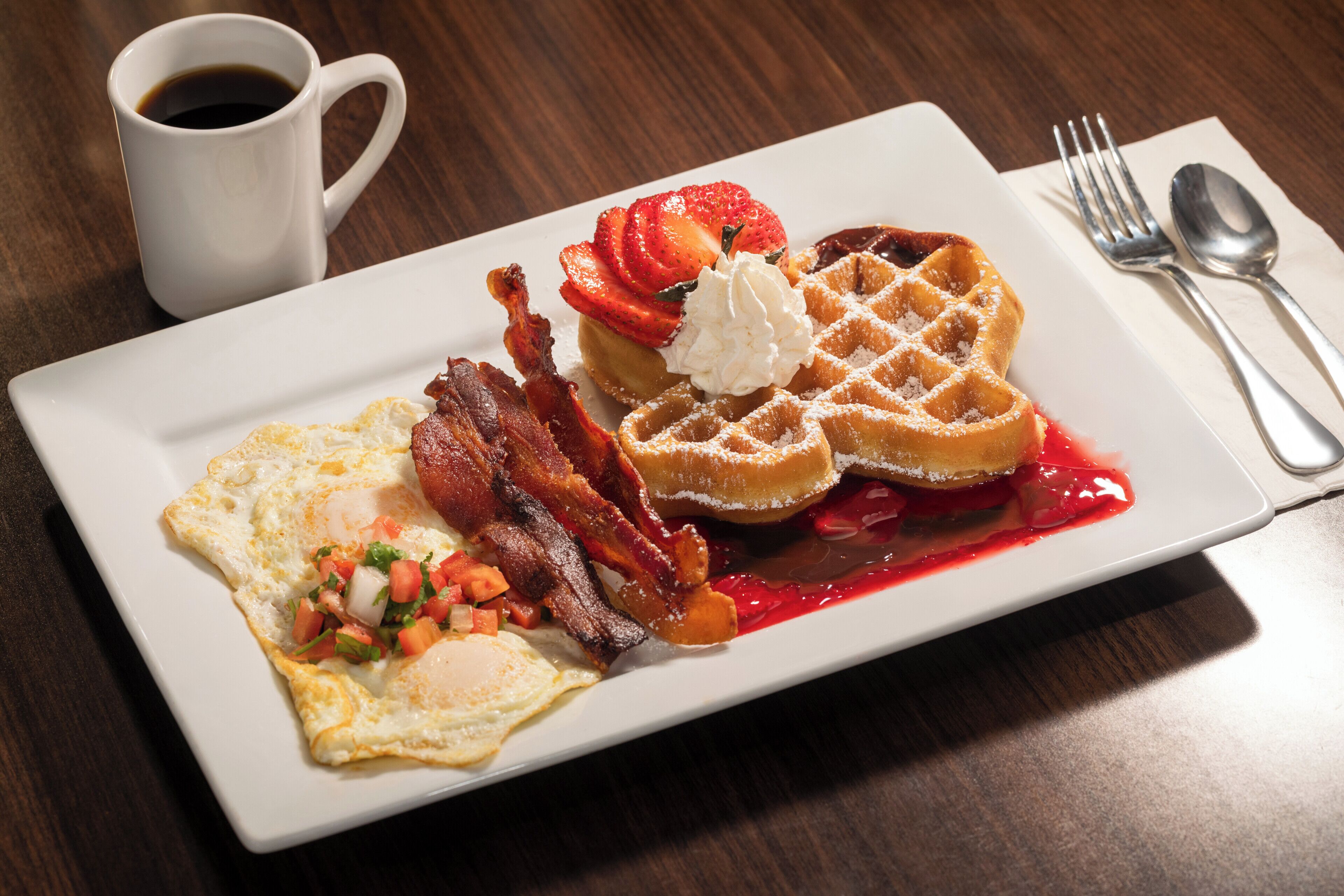 Une assiette de petit-déjeuner comprenant des gaufres, des fraises, de la crème fouettée, du bacon et un œuf au plat.