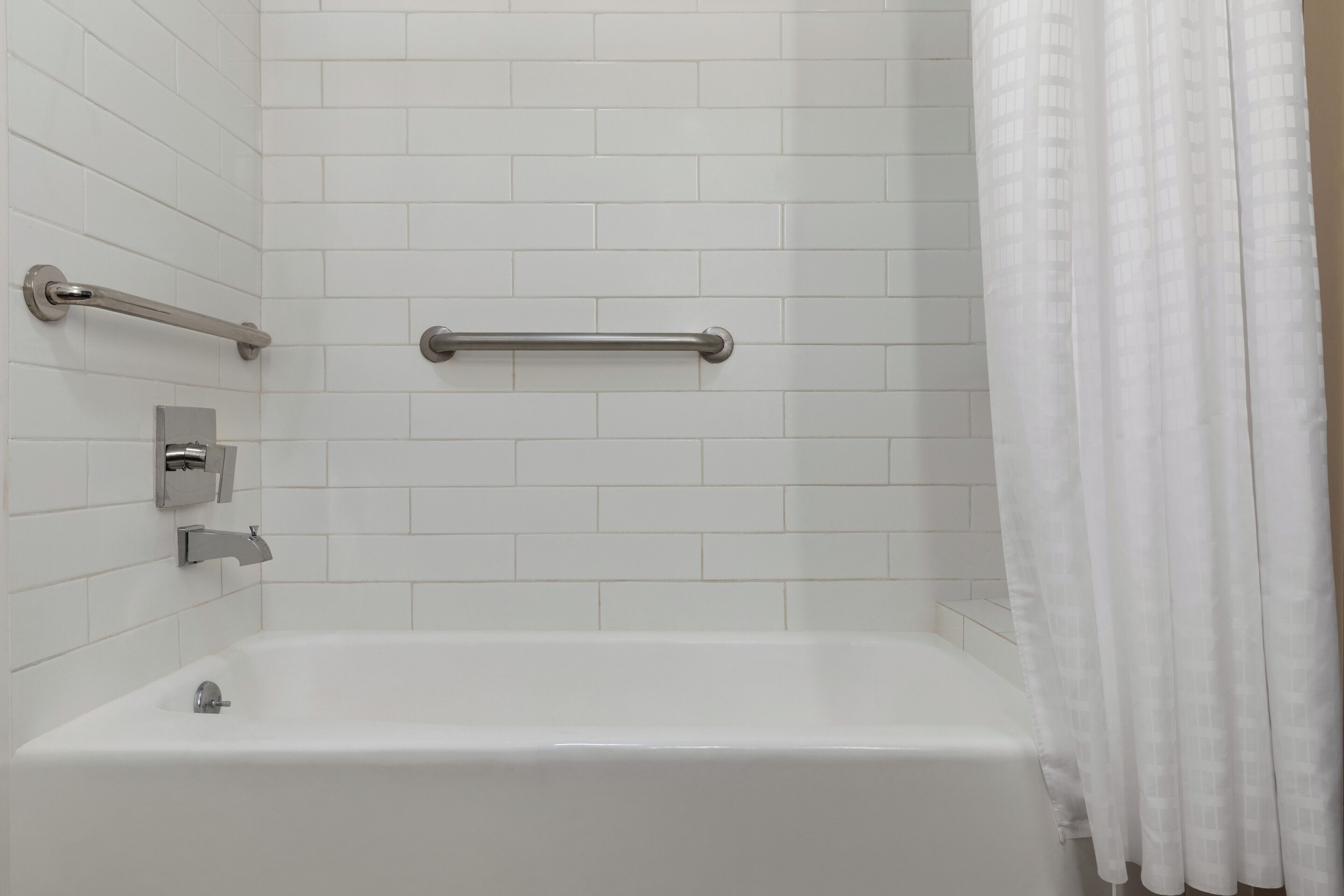 A bathroom with white tiles, a bathtub, a handrail, and a shower curtain.