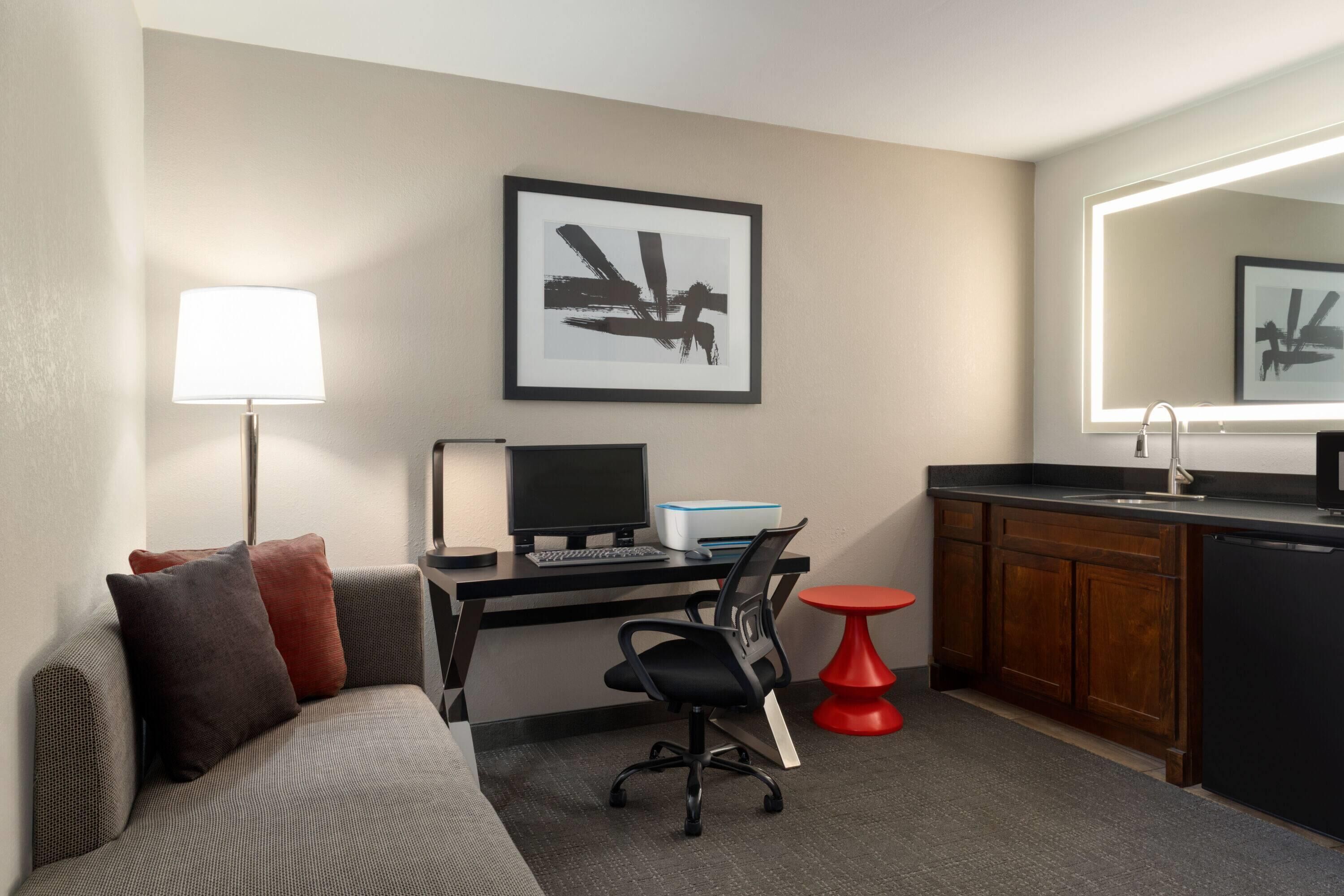 A compact hotel room with a bed, a desk with a computer, a chair, a small red table, a kitchen area with a sink, and a framed artwork on the wall.