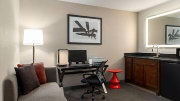 A compact hotel room with a bed, a desk with a computer, a chair, a small red table, a kitchen area with a sink, and a framed artwork on the wall.