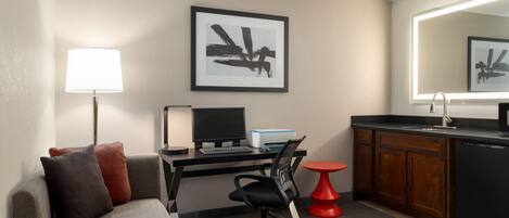 Une chambre d’hôtel compacte comprenant un lit, un bureau avec un ordinateur, une chaise, une petite table rouge, un coin cuisine avec un évier et une œuvre d’art encadrée au mur.
