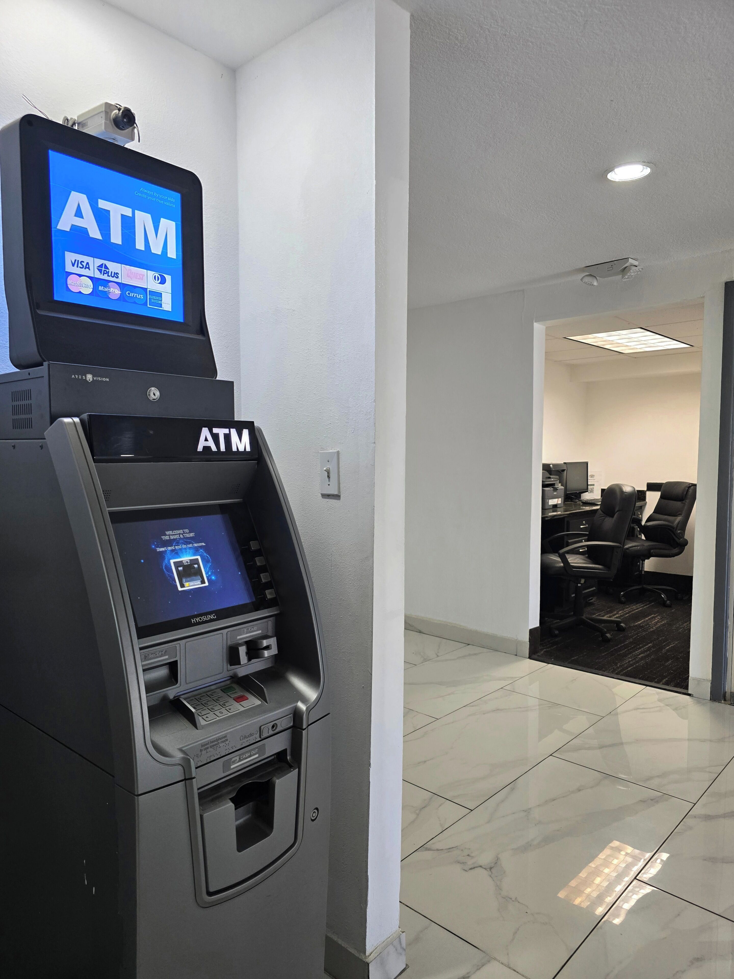 Un distributeur automatique de billets doté d’un écran numérique, un espace d’accueil avec un comptoir et des chaises, et un sol carrelé.
