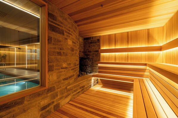 Una sauna con bancos de madera y pared de piedra, una puerta de cristal que da a una zona de piscina y techo de madera.
