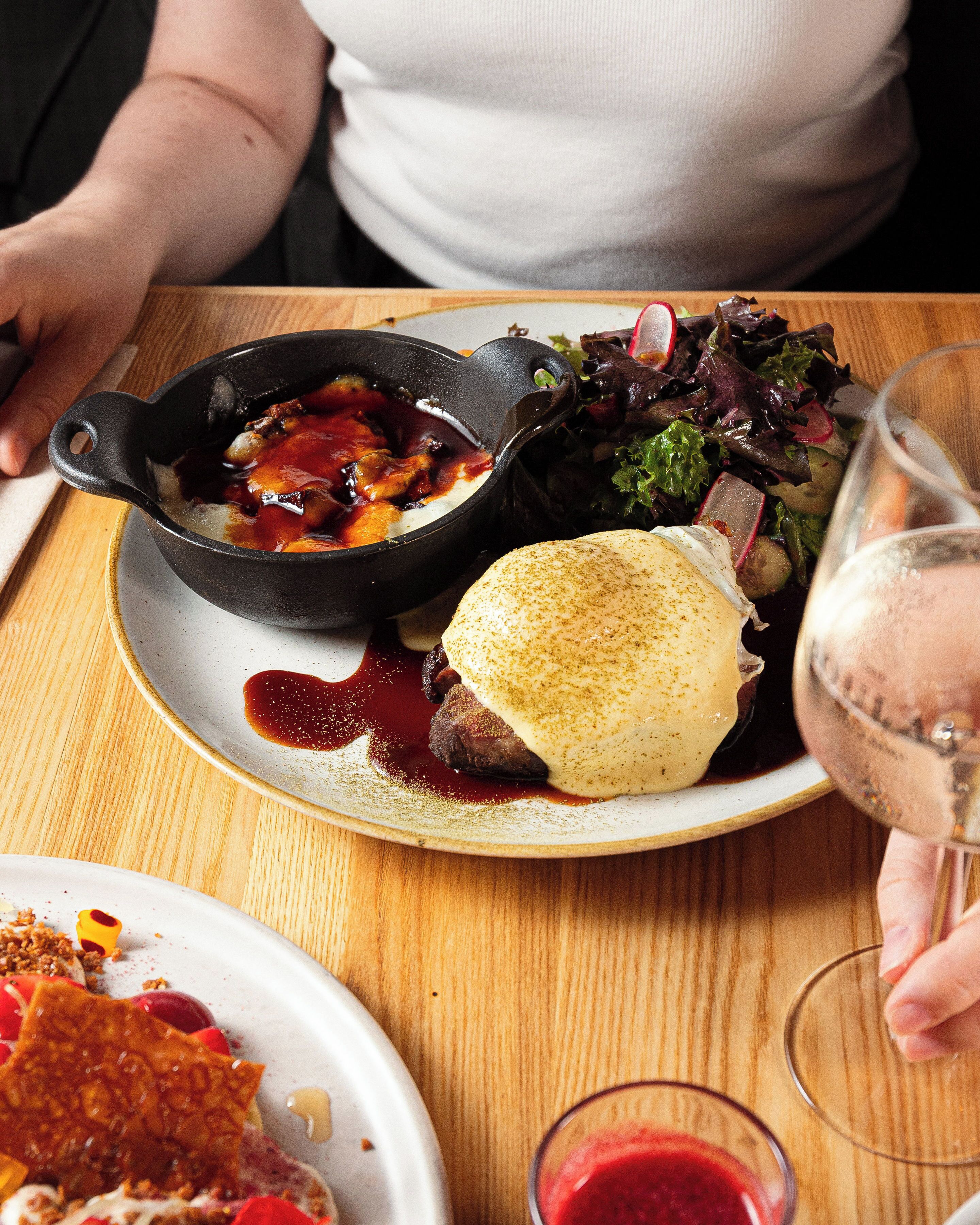 A dining table with a plate of food, a glass of red wine, and a person holding a fork.