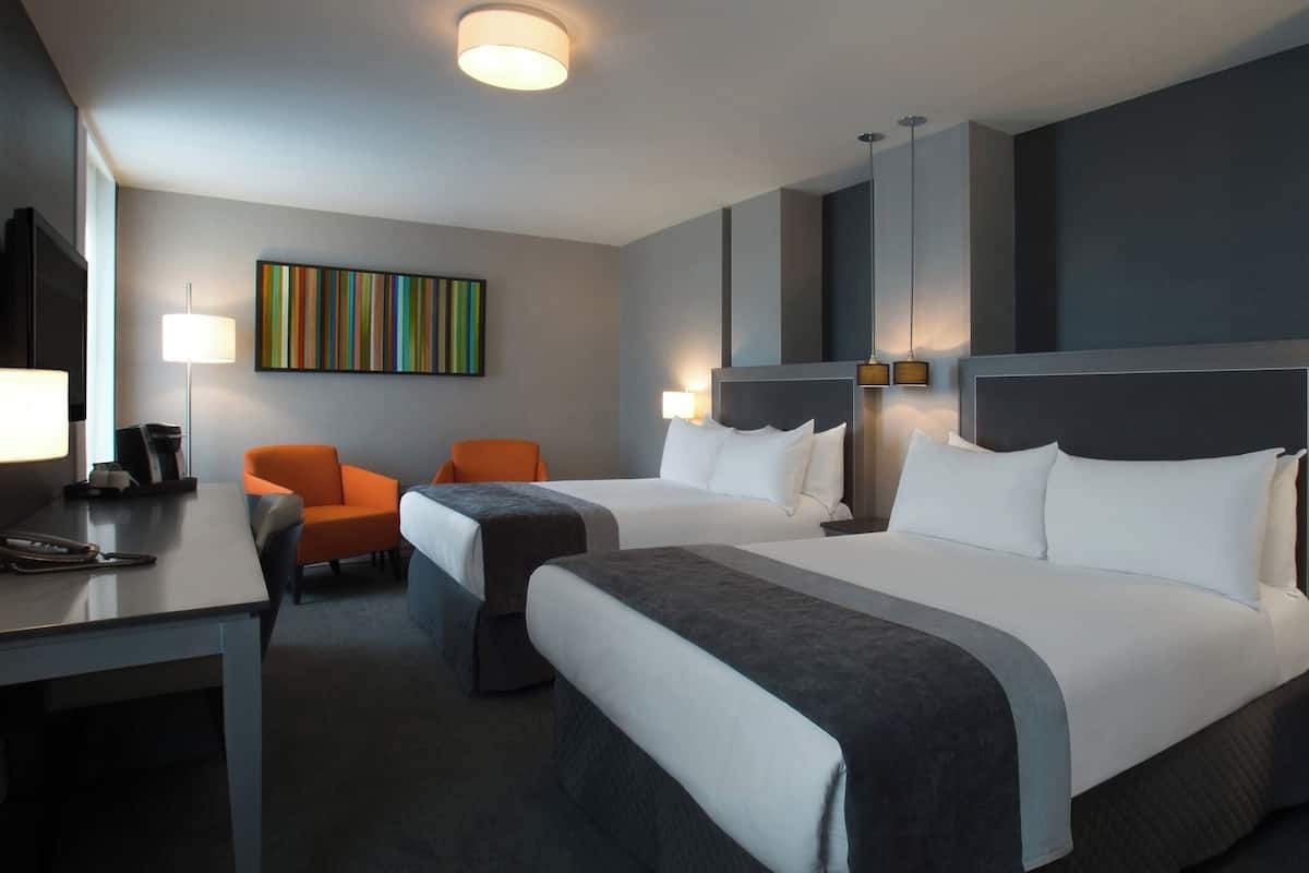 Une chambre d’hôtel avec deux lits, un bureau avec un téléphone, deux chaises, une machine à café et un tableau abstrait aux couleurs vives accroché au mur.