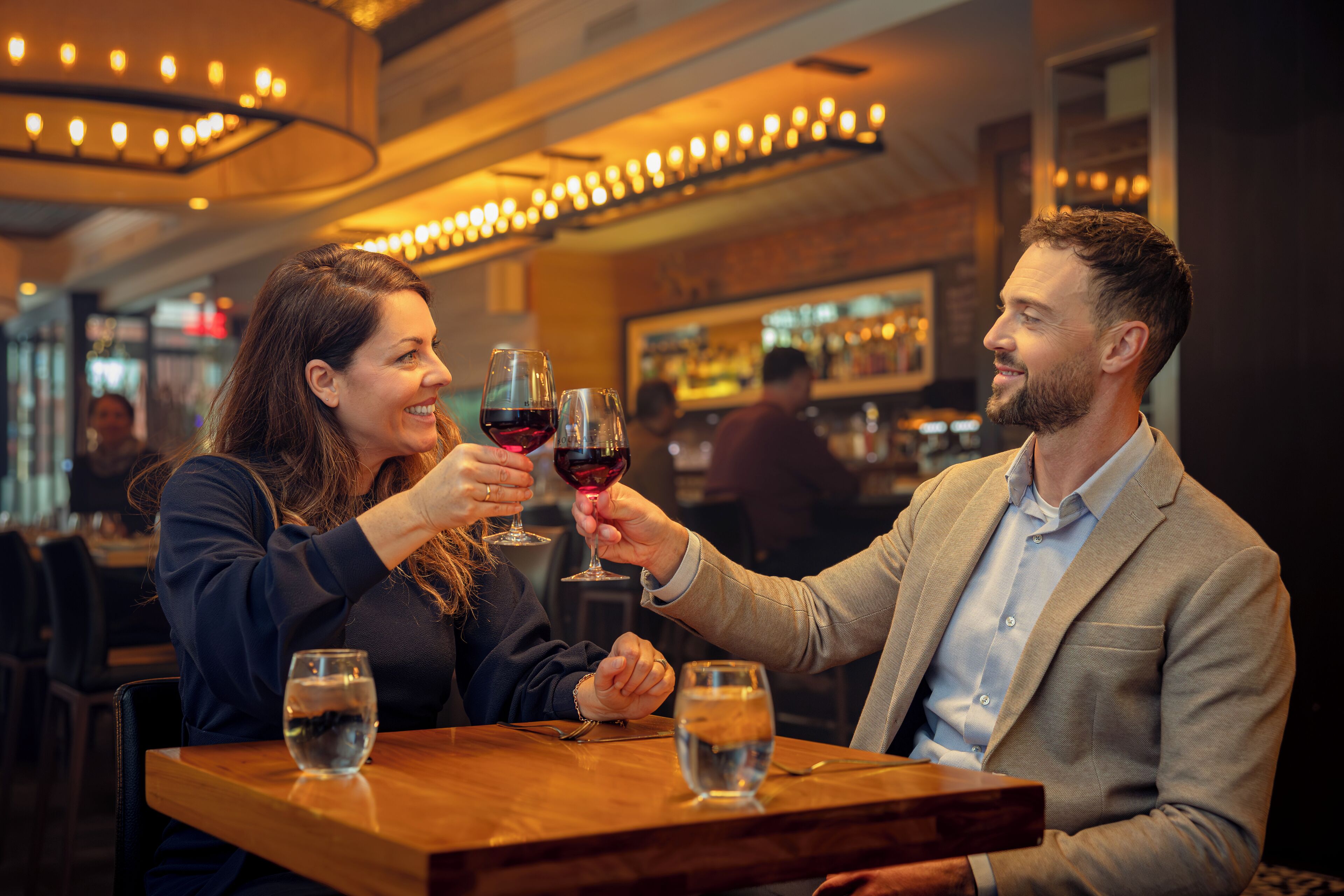 Dos personas brindando con copas de vino en una mesa de madera en un restaurante con luz cálida y un bar al fondo.