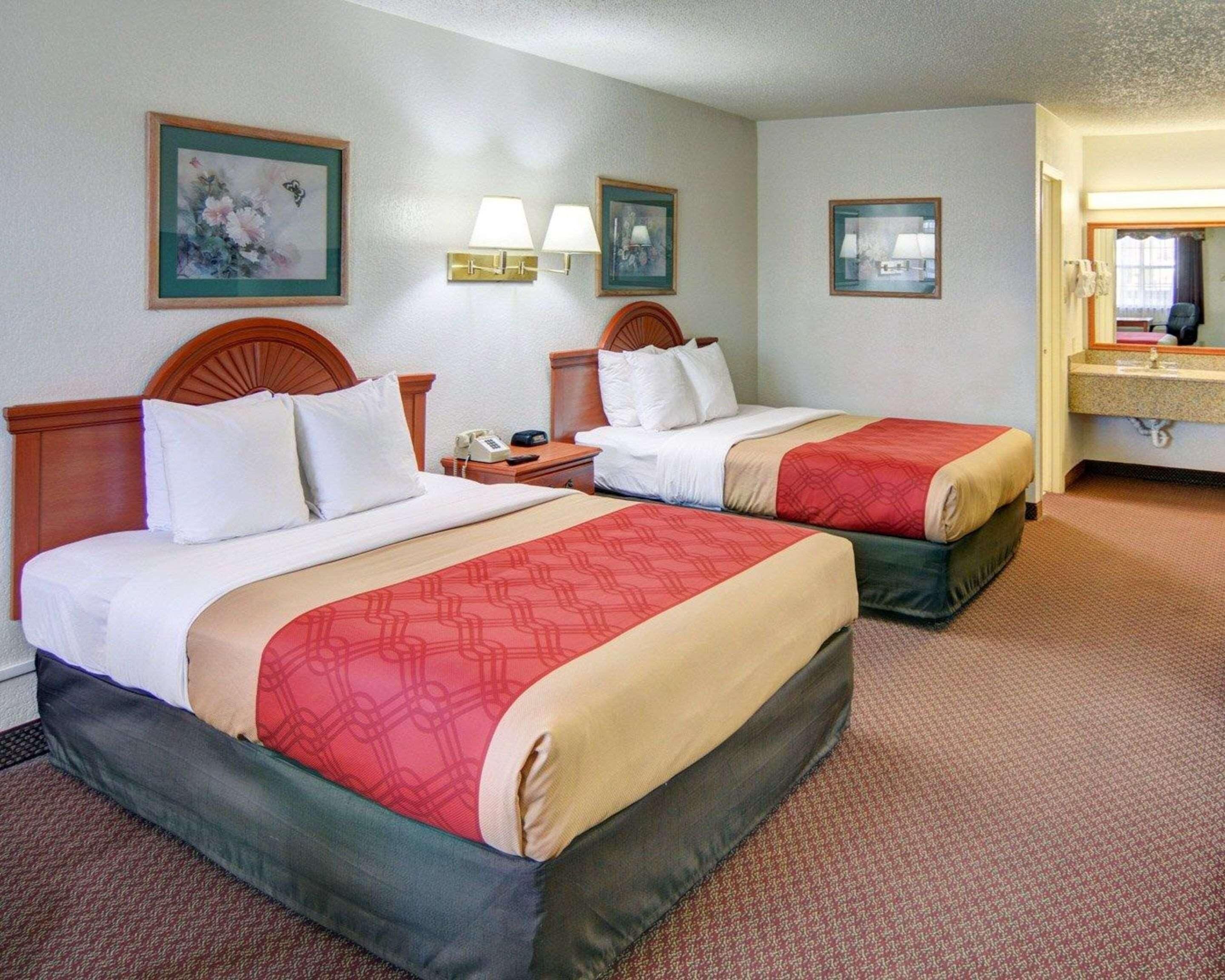 Habitación de hotel con dos camas, una mesita de noche, una lámpara, un cuadro enmarco y un baño con lavamanos y espejo.