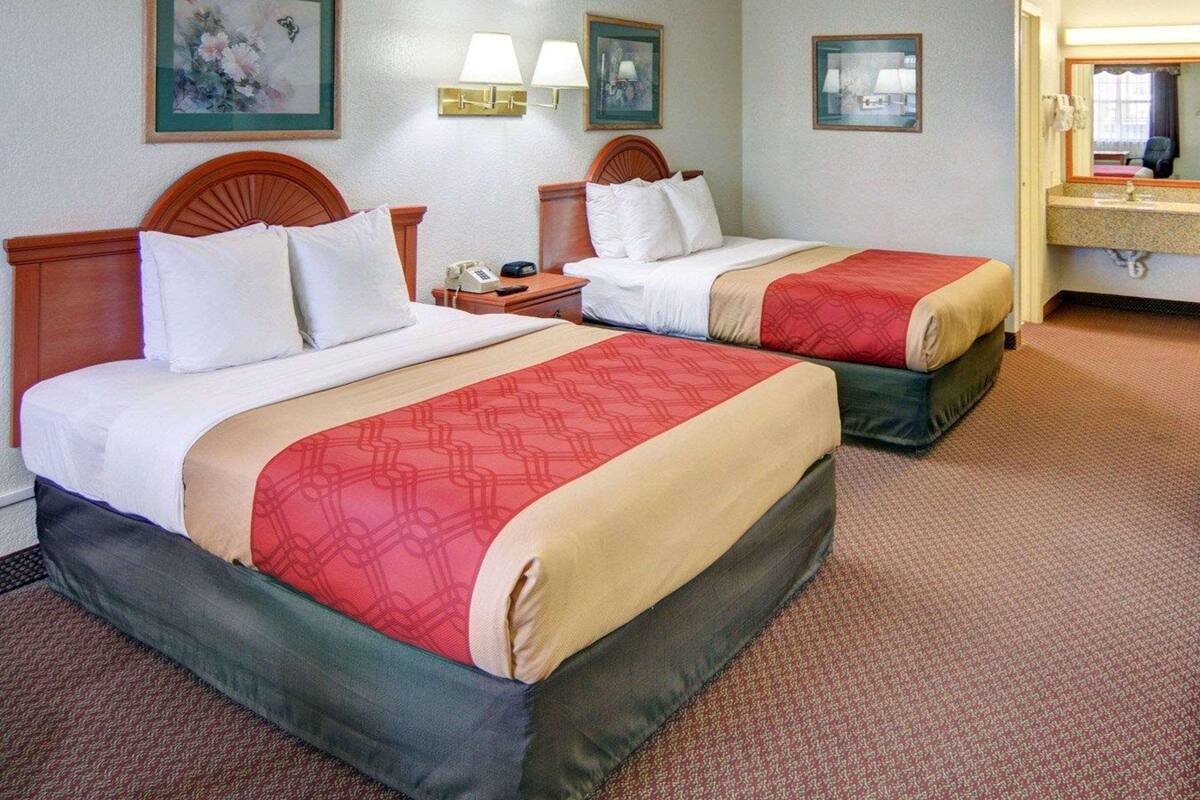 A hotel room with two beds, a nightstand, a lamp, a framed picture, and a bathroom with a sink and mirror.