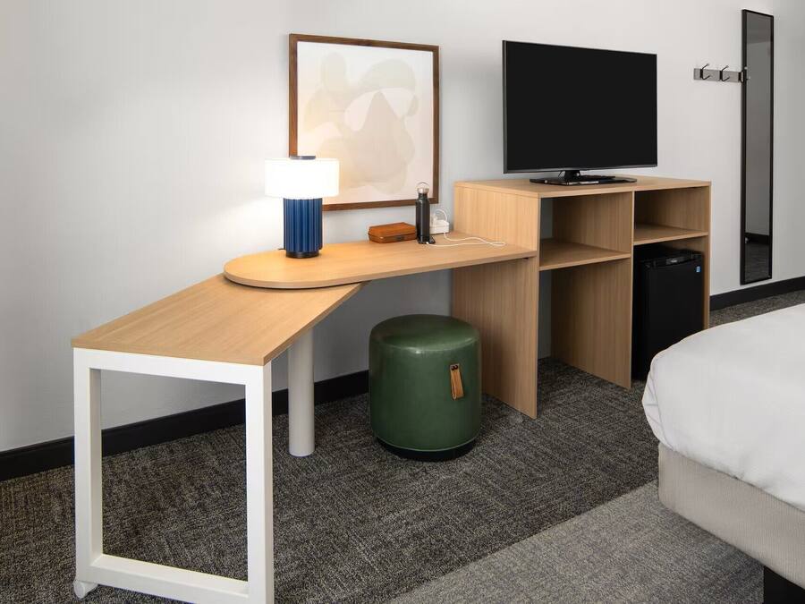 Quarto de hotel com uma escrivaninha, televisão de tela plana, uma cama e um cesto de lixo verde.