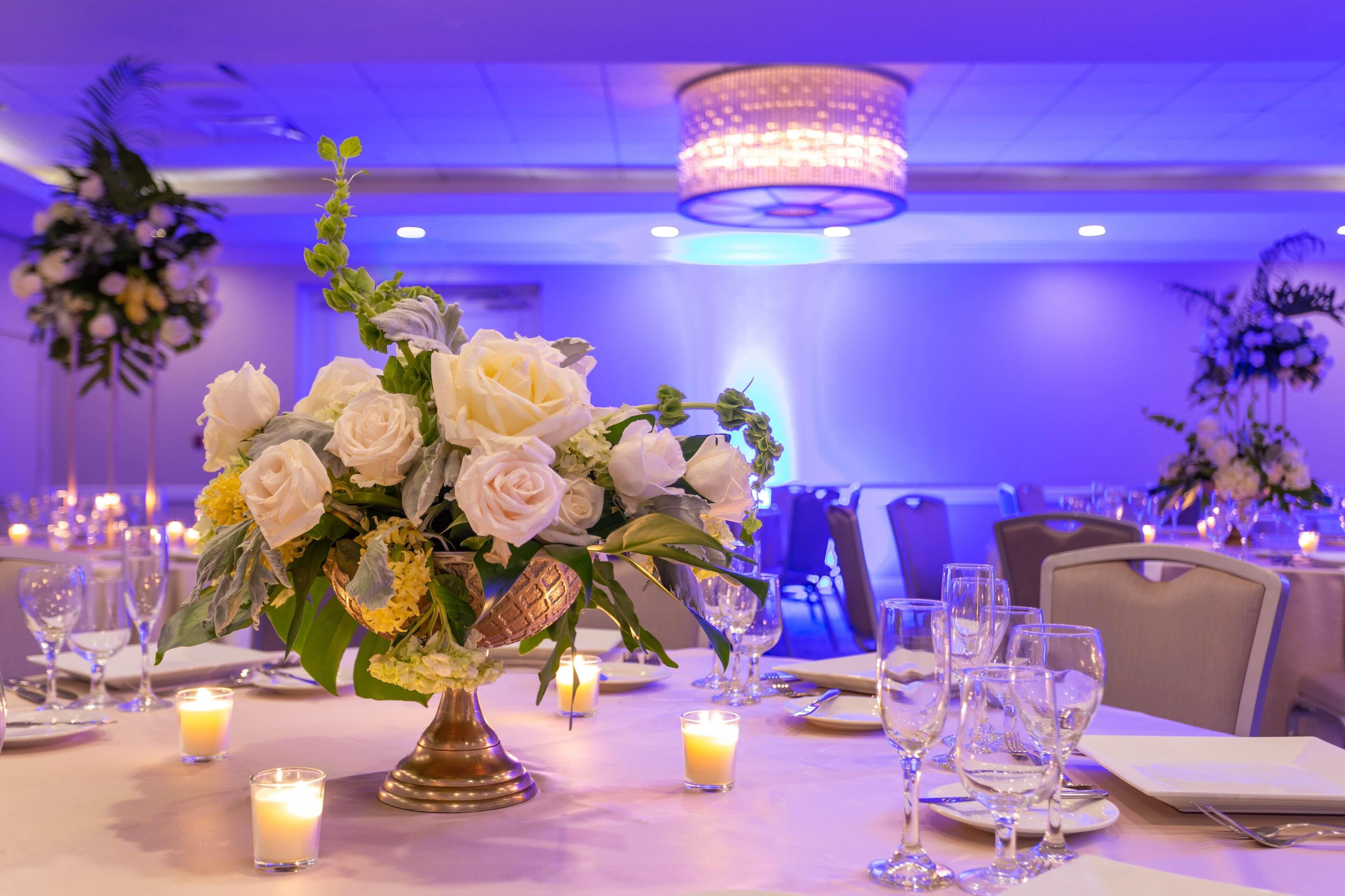Salão de banquetes com mesas montadas para refeições, com um arranjo de rosas brancas e flores amarelas em uma tigela decorativa.