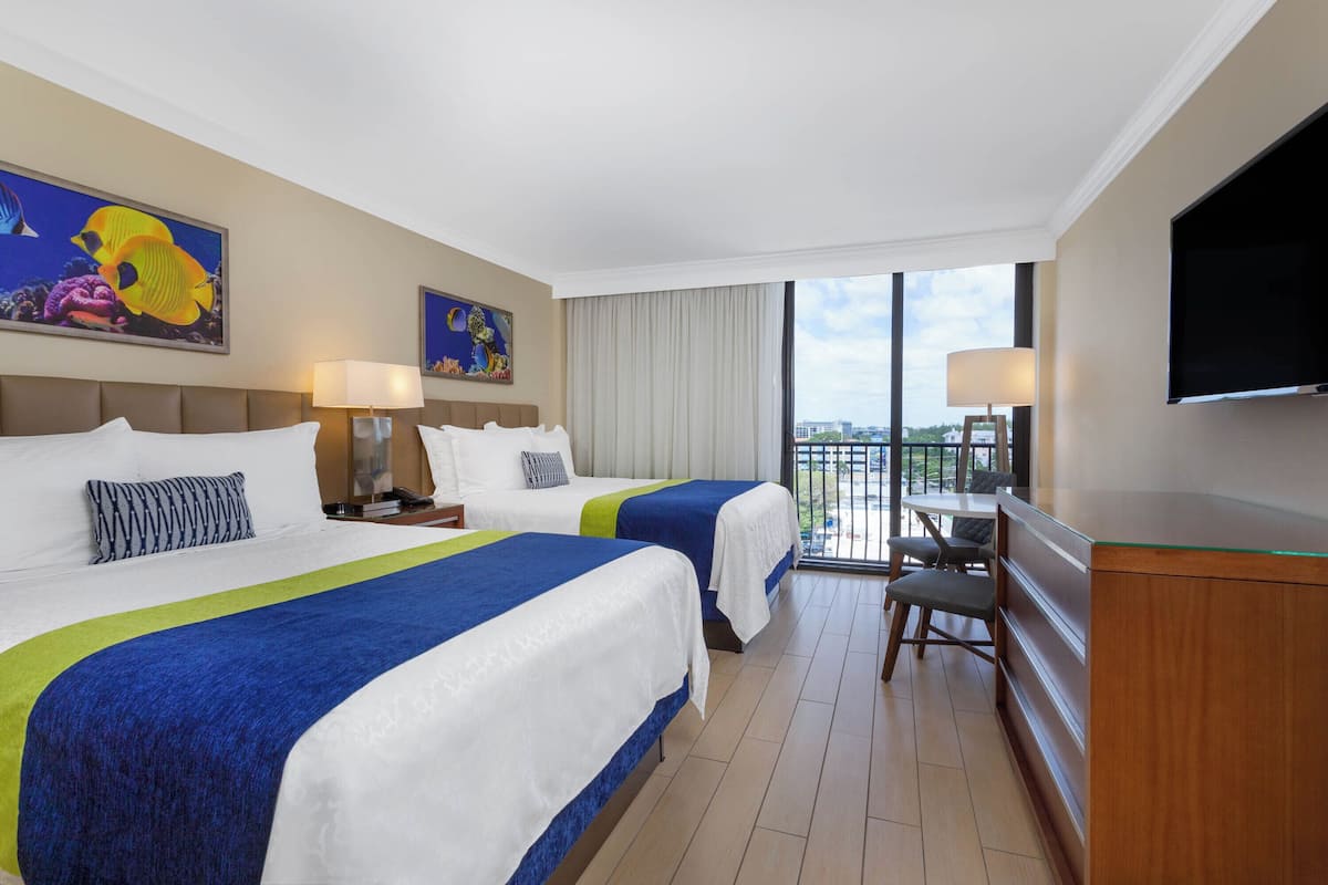Habitación de hotel con dos camas, un escritorio, un televisor y un balcón con vista a la ciudad.