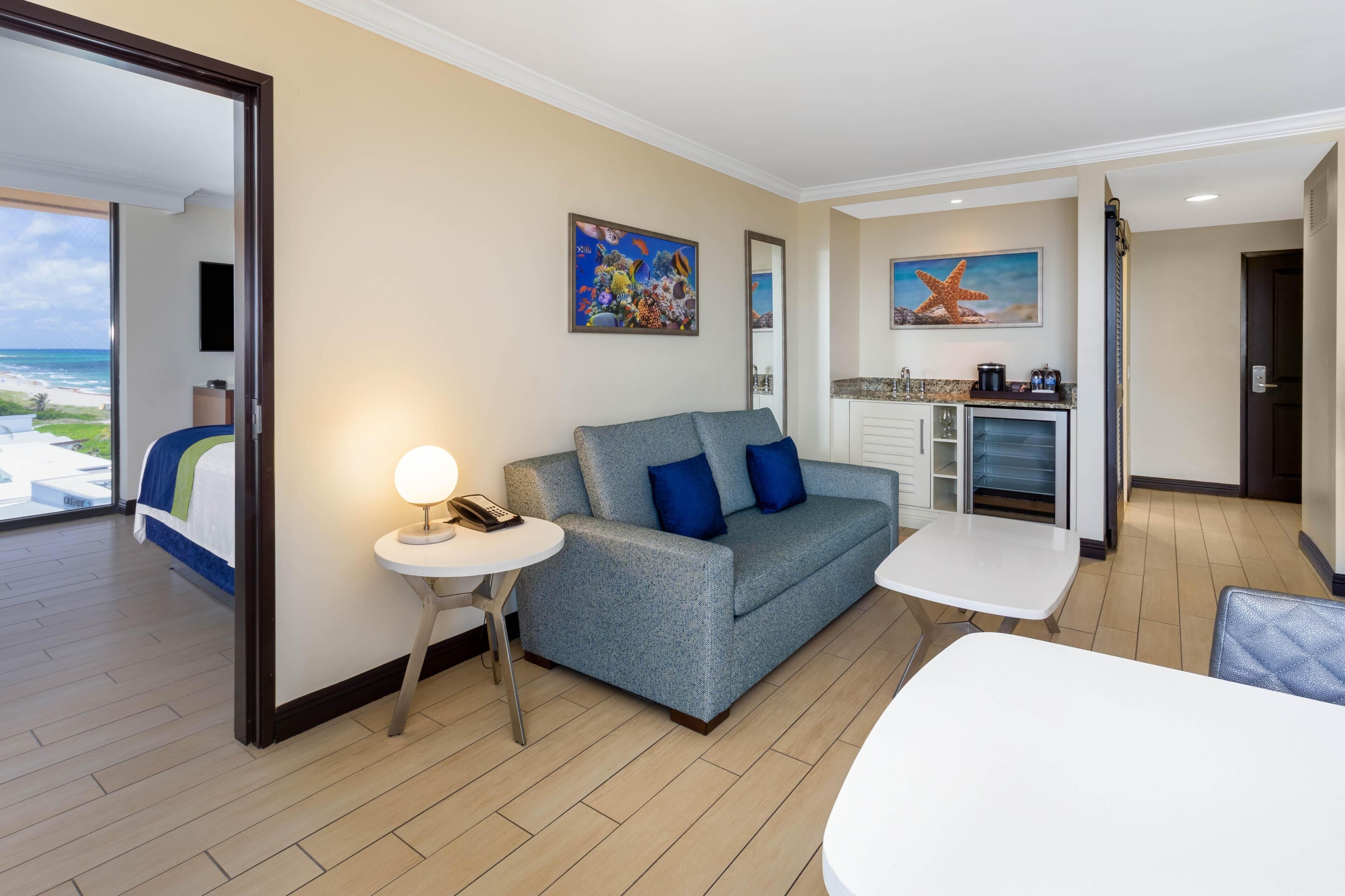 Ein modernes Wohnzimmer mit einem grauen Sofa, einem kleinen runden Tisch mit Lampe und Blick auf das Meer durch ein großes Fenster.