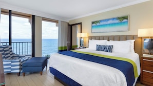 Habitación de hotel con una cama grande, vistas al océano, un balcón con una silla y un cuadro con una playa.