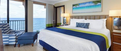 Une chambre d’hôtel avec un grand lit, une vue sur l’océan, un balcon avec une chaise et un tableau représentant une scène de plage.