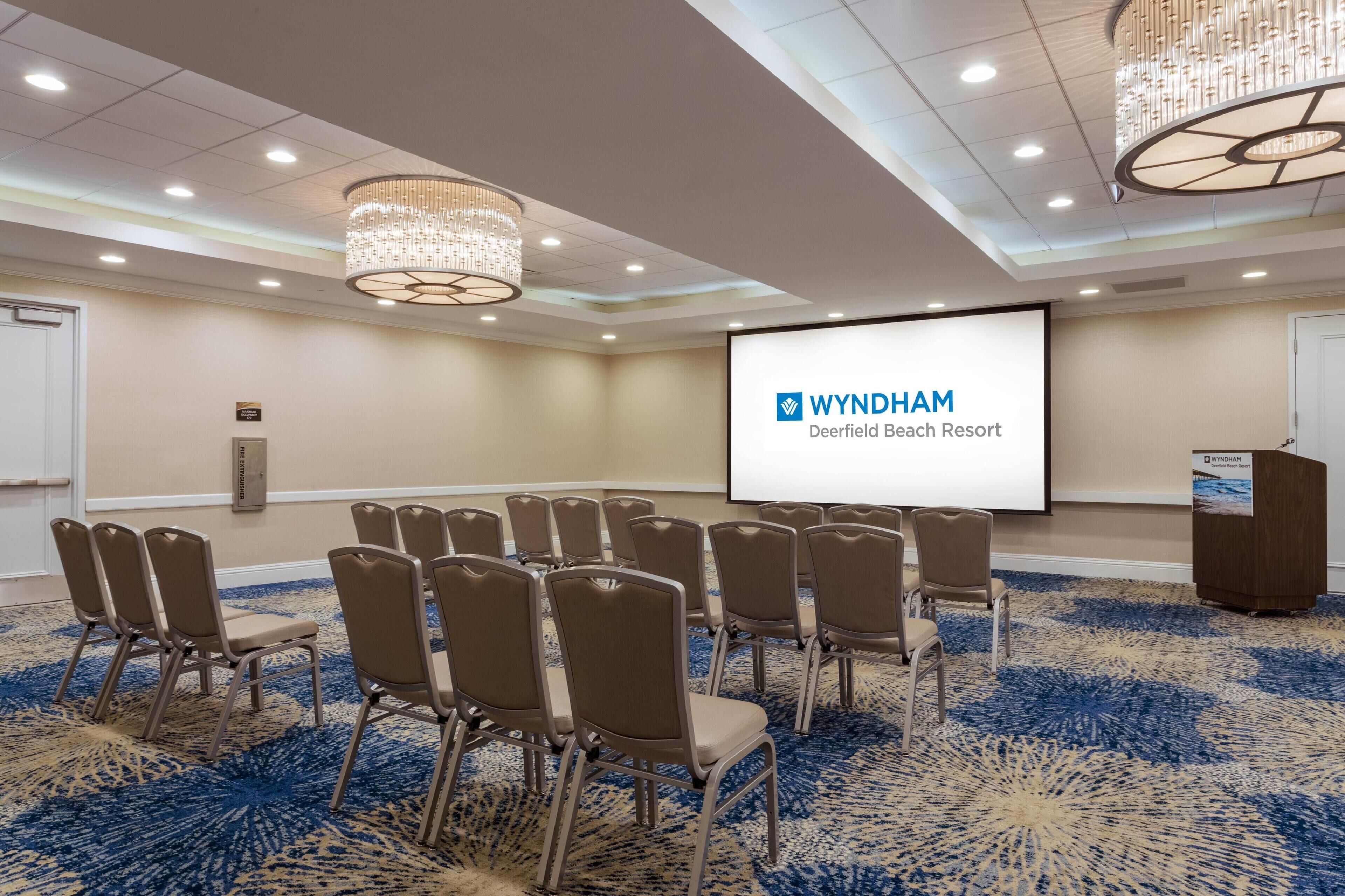 A conference room with a projector screen displaying 'WYNDHAM Deerfield Beach Resort', rows of chairs, a podium, and a chandelier.