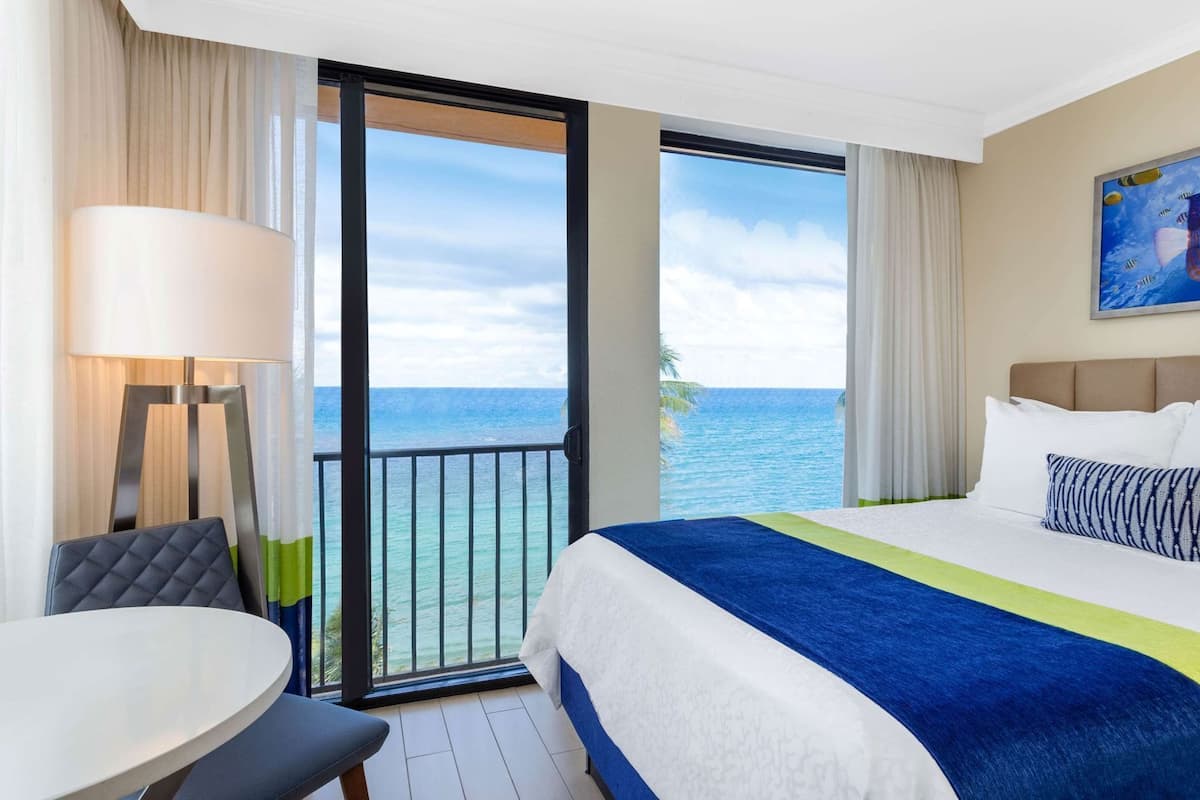 Habitación de hotel con cama, una mesa, una silla, una lámpara, un cuadro y vistas al océano.