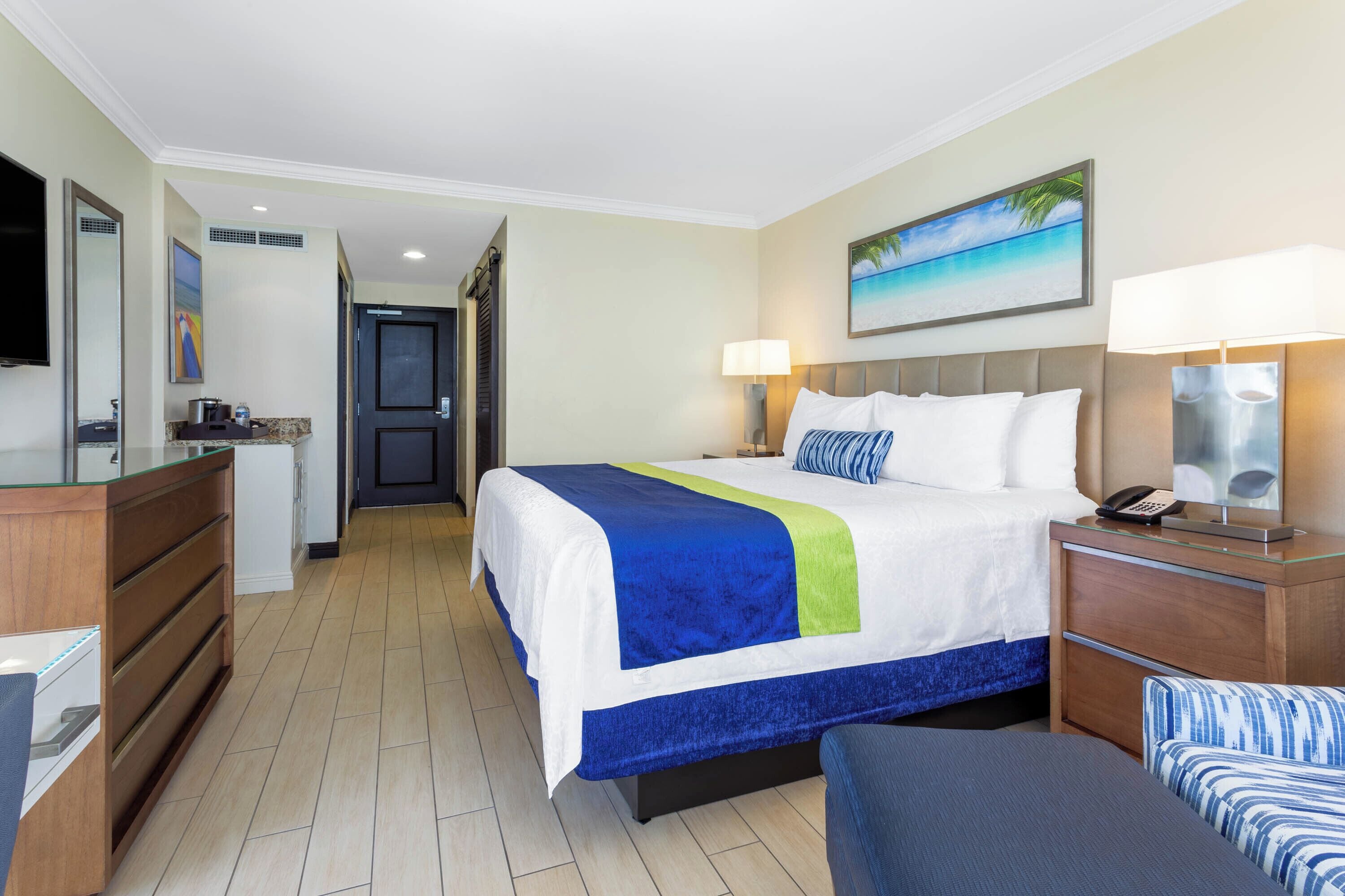 Habitación de hotel con cama, lámparas de noche, escritorio, silla, televisor y un cuadro de una playa.