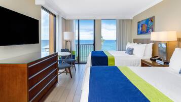 Habitación de hotel con dos camas, televisión, escritorio y balcón con vista al mar.