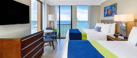 Habitación de hotel con dos camas, televisión, escritorio y balcón con vista al mar.