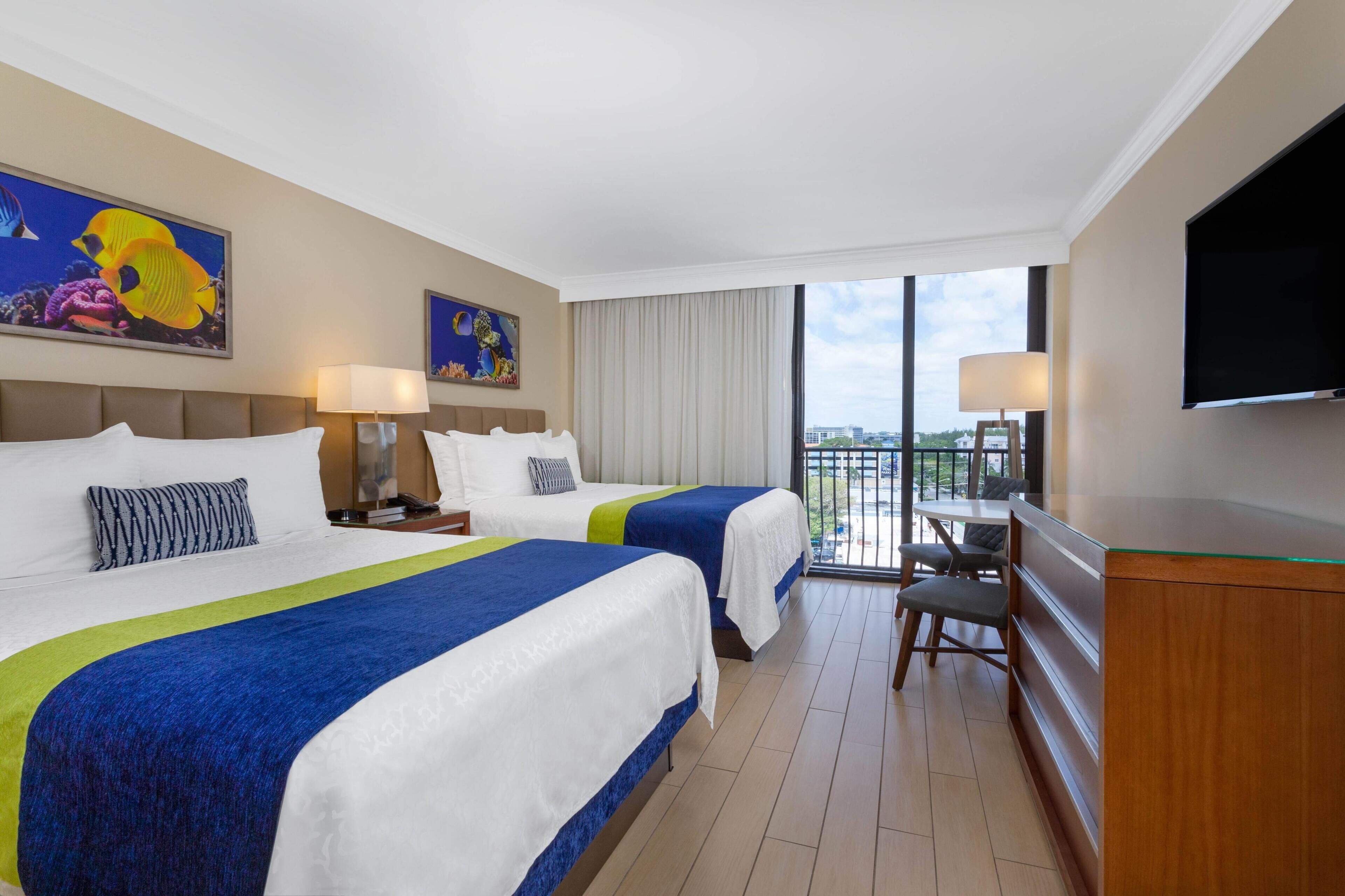 A hotel room with two beds, a desk, a TV, and a balcony with a city view.