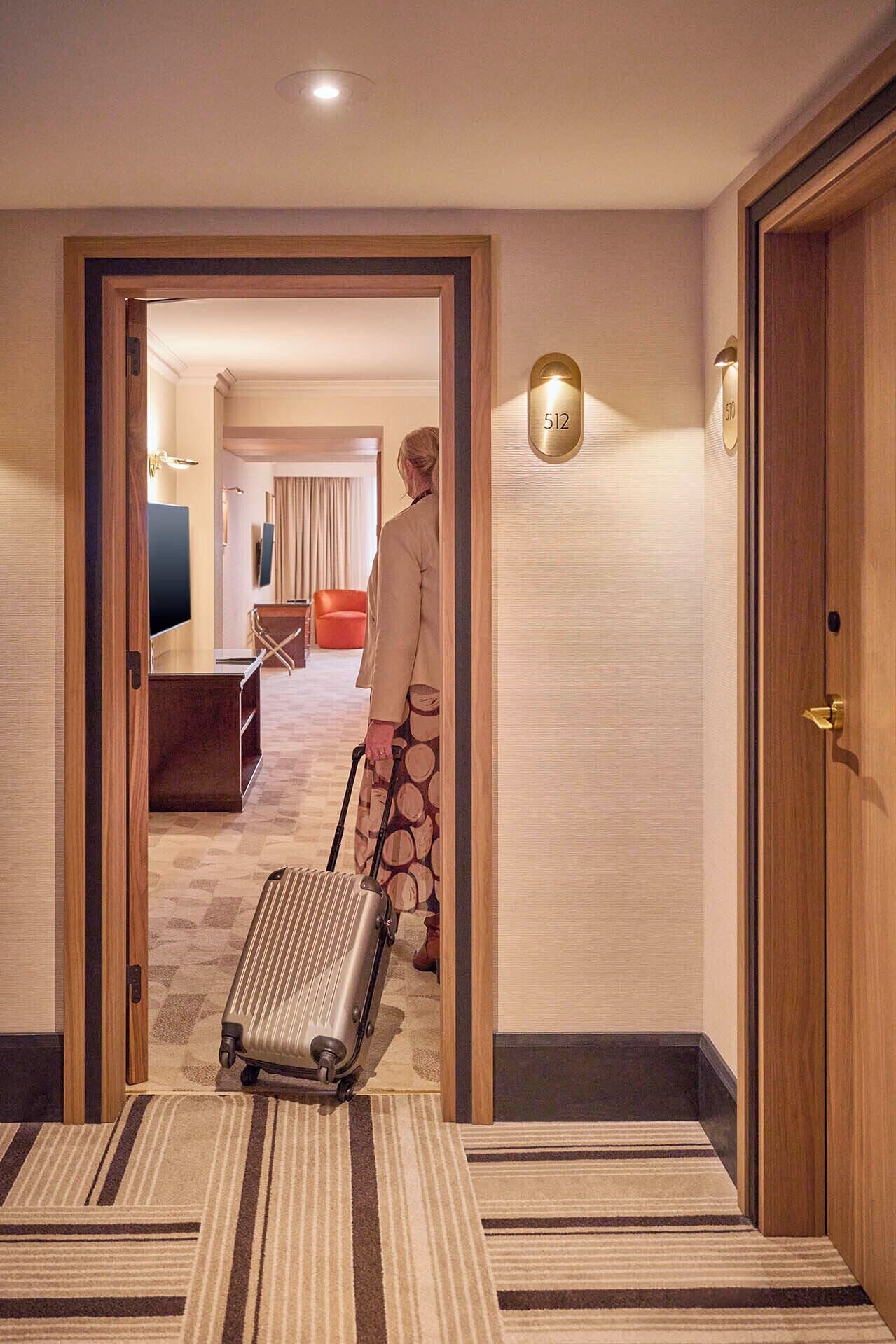 A hotel hallway with a wooden door, a person pulling a suitcase, and a room visible in the background.