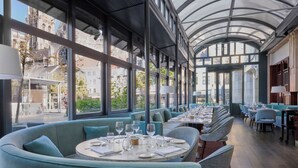 Un ristorante con un lungo tavolo e una zona salotto, ampie finestre e vista su un edificio storico.