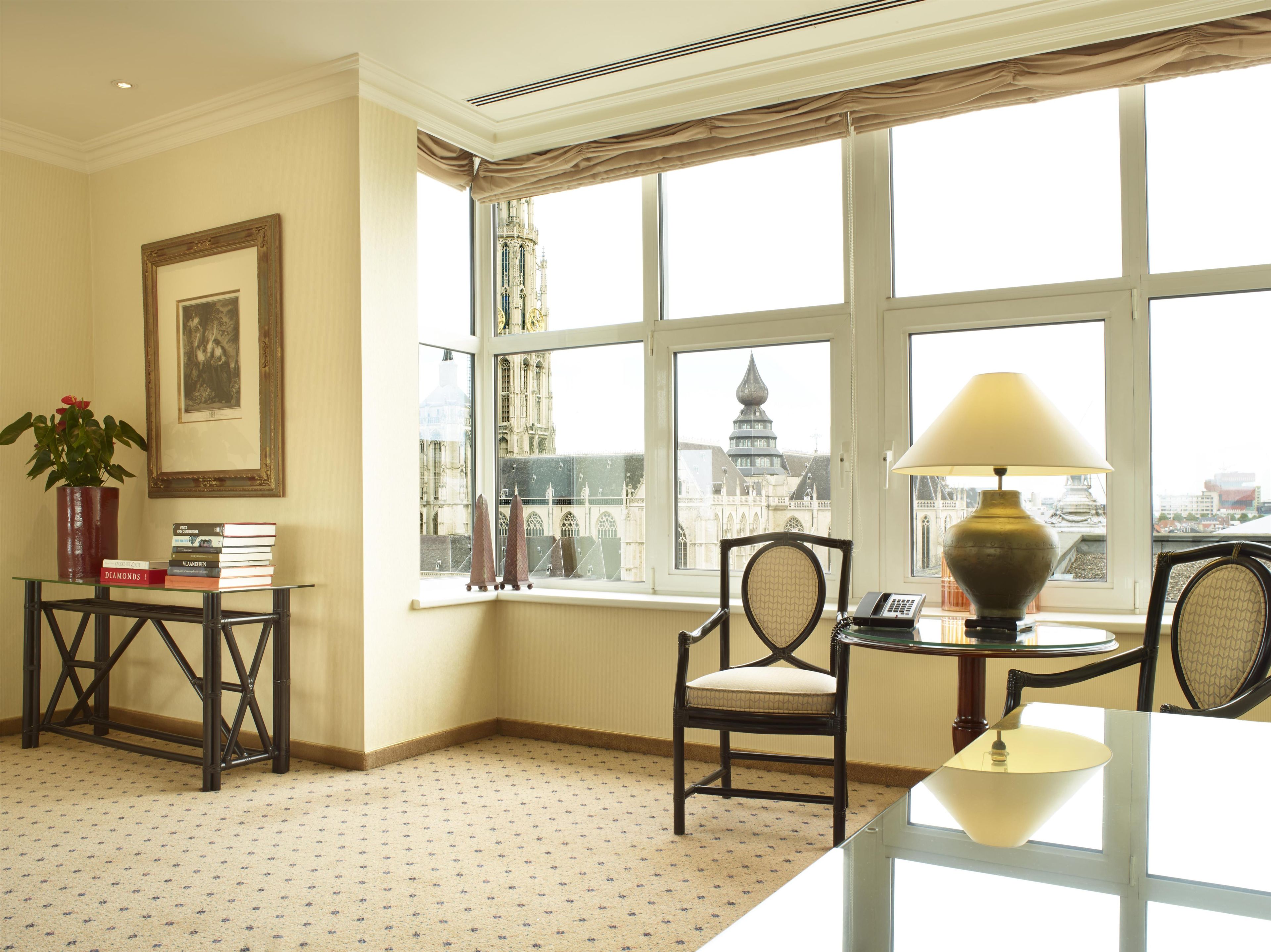 A room with a large window, a table with a lamp, a chair, a small table with books and a plant, and a framed picture on the wall.