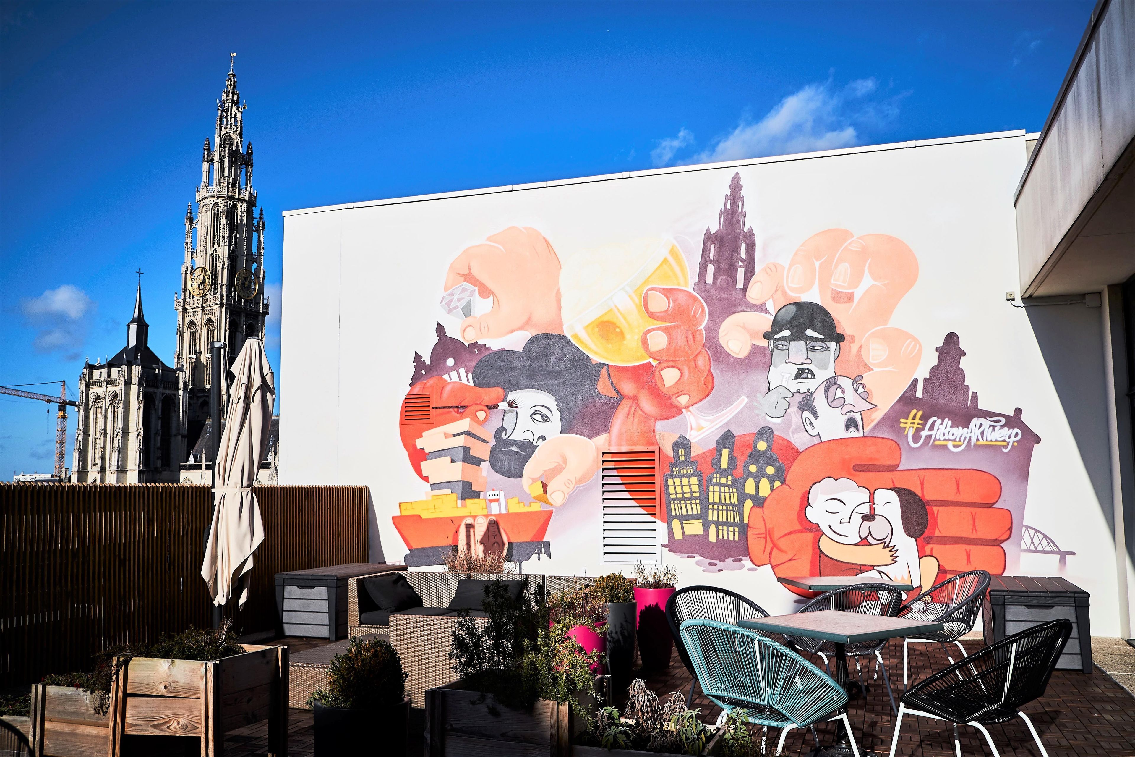 Un mural con personajes de dibujos animados y un hashtag, un edificio con aguja, y asientos exteriores con plantas.