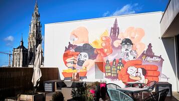 Un mural con personajes de dibujos animados y un hashtag, un edificio con aguja, y asientos exteriores con plantas.