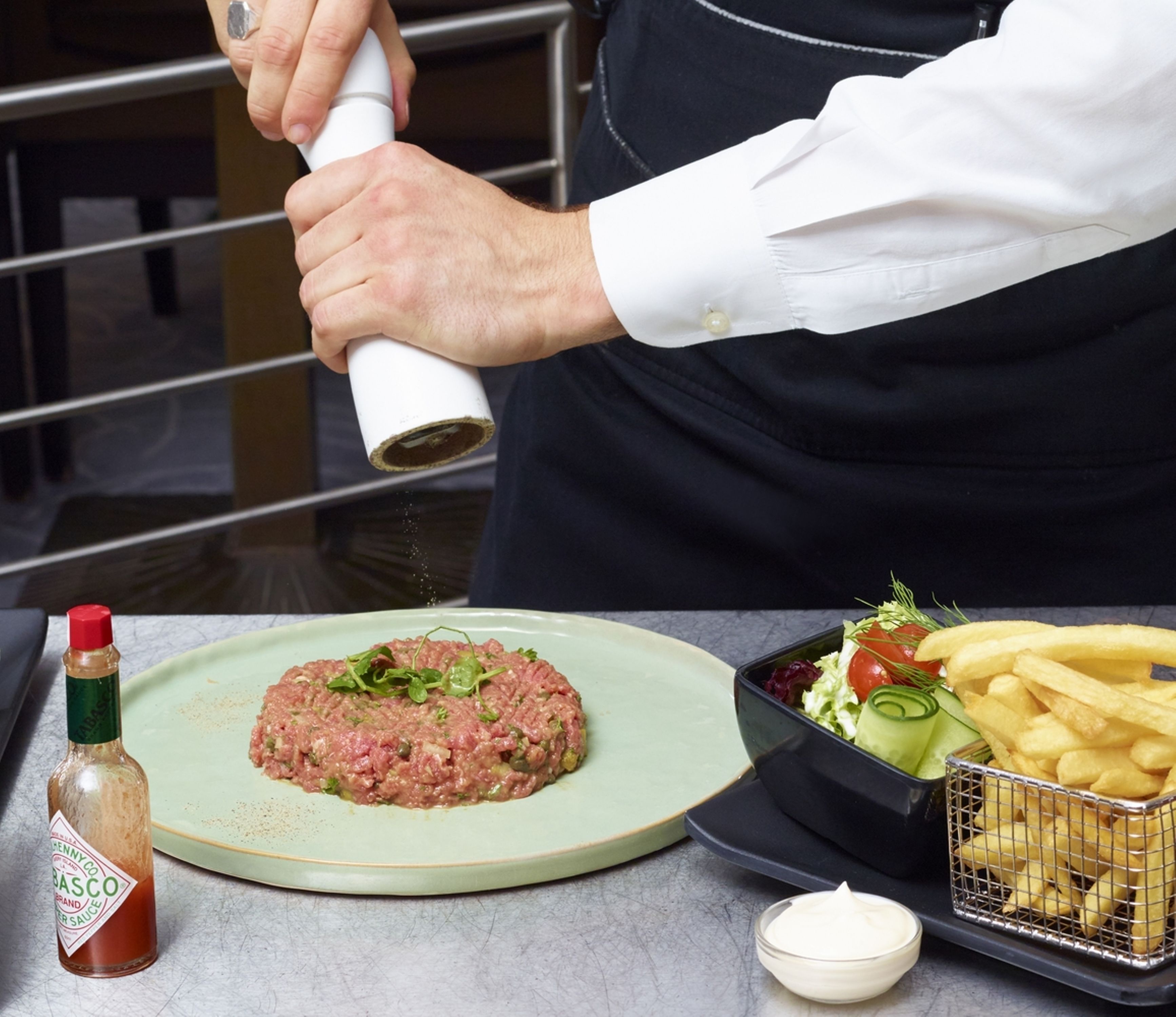 A chef preparing steak tartare on a plate, accompanied by a salad and fries.
