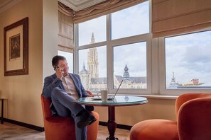 Un hombre sentado junto a una ventana con vistas a un edificio histórico, una laptop sobre una mesa de vidrio y un cuadro enmarcado en la pared.