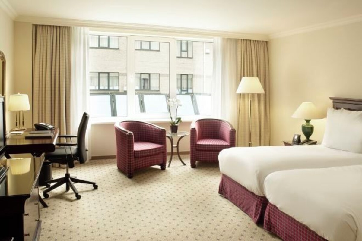 A hotel room with a bed, two armchairs, a desk with a lamp, a chair, and a window with curtains.