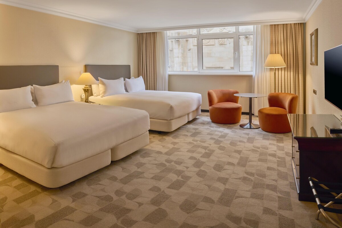 Habitación de hotel con dos camas, una mesita, dos sillones, un televisor y una ventana con cortinas.