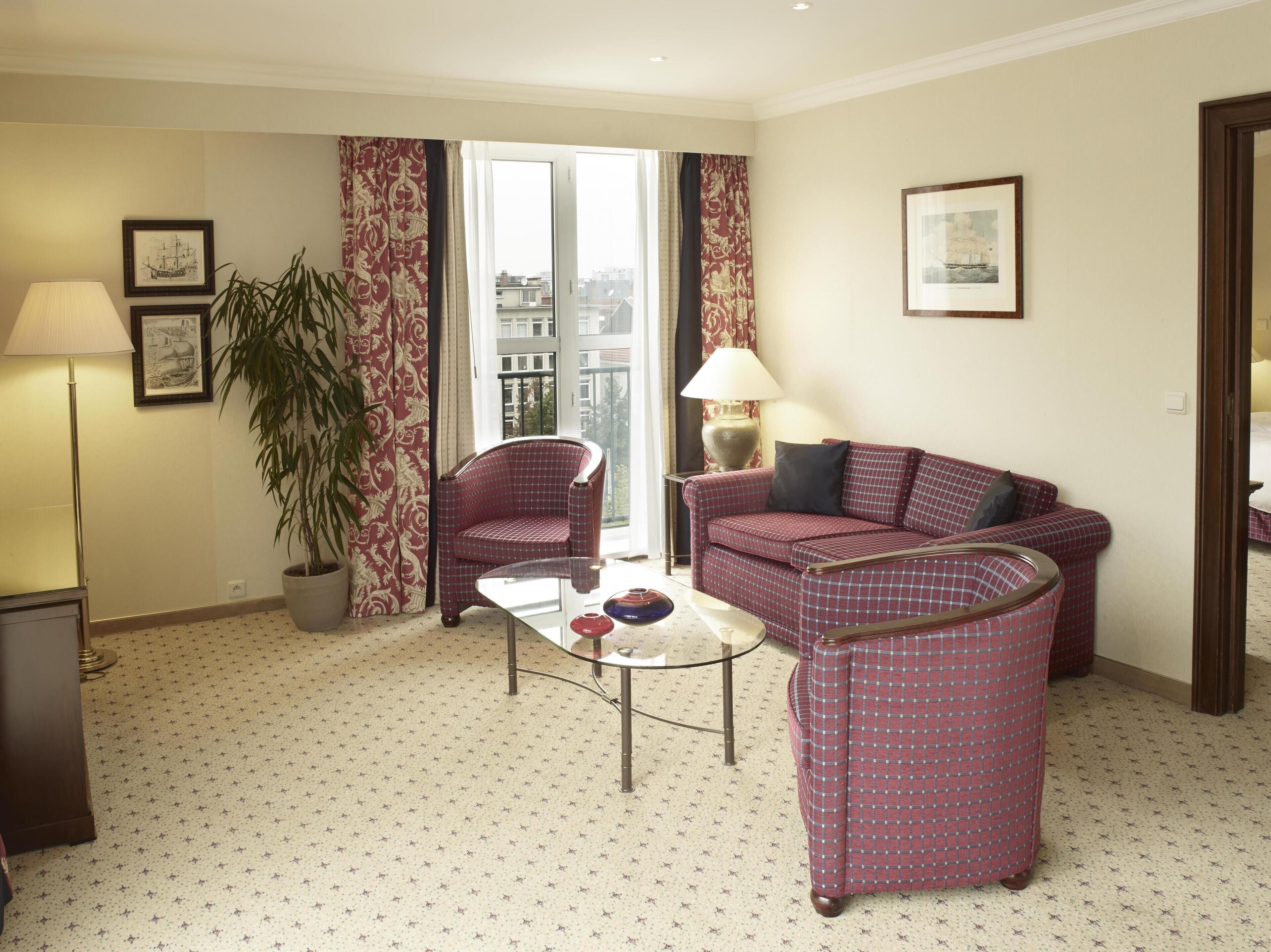 A hotel room with a checkered sofa, a small round table, a lamp, a potted plant, and framed pictures on the walls.