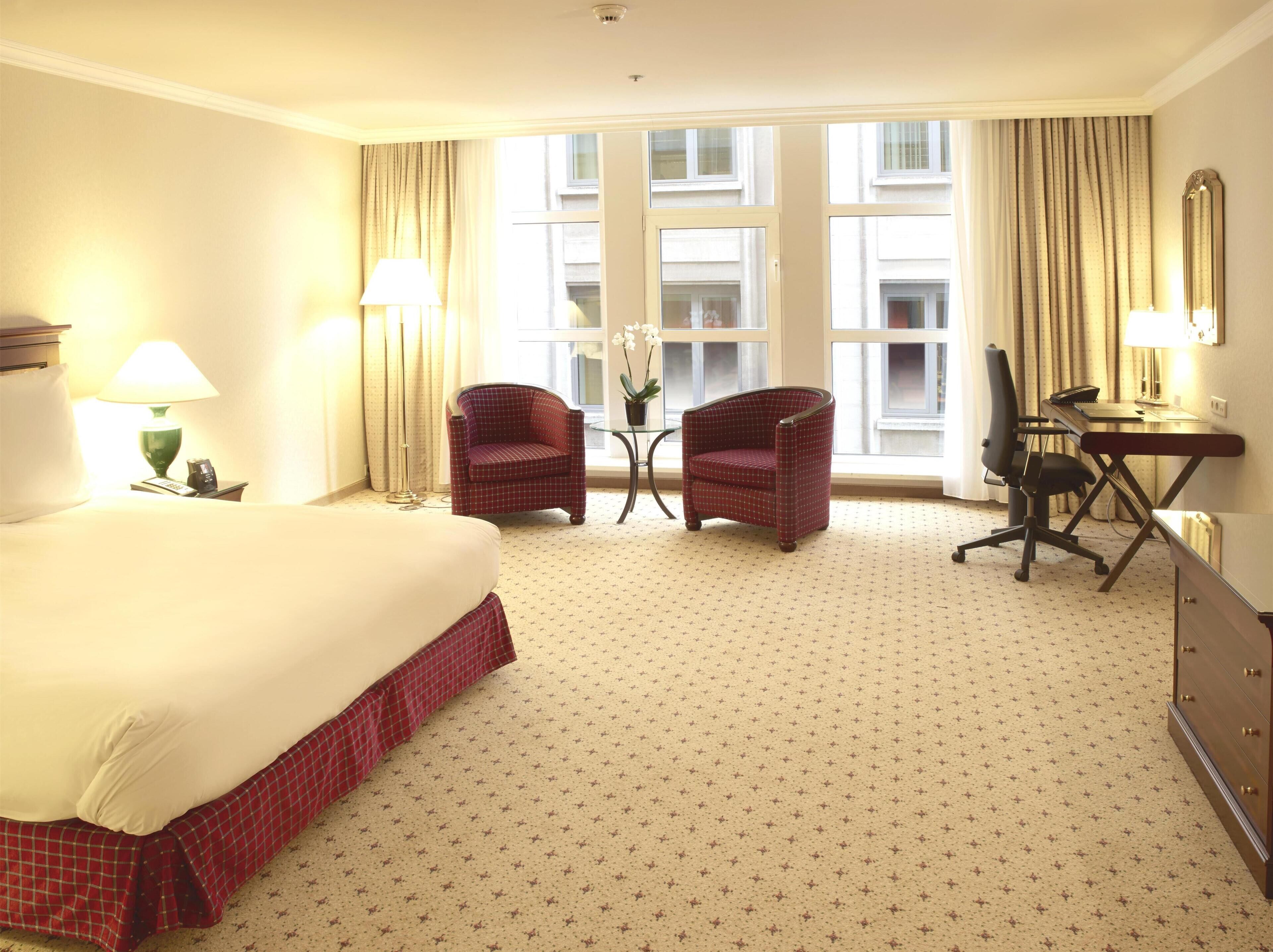 A hotel room with a large bed, two armchairs, a desk with a chair, and a view of buildings through the windows.