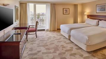 A hotel room with two beds, a desk, a chair, a TV, and a large window with curtains.
