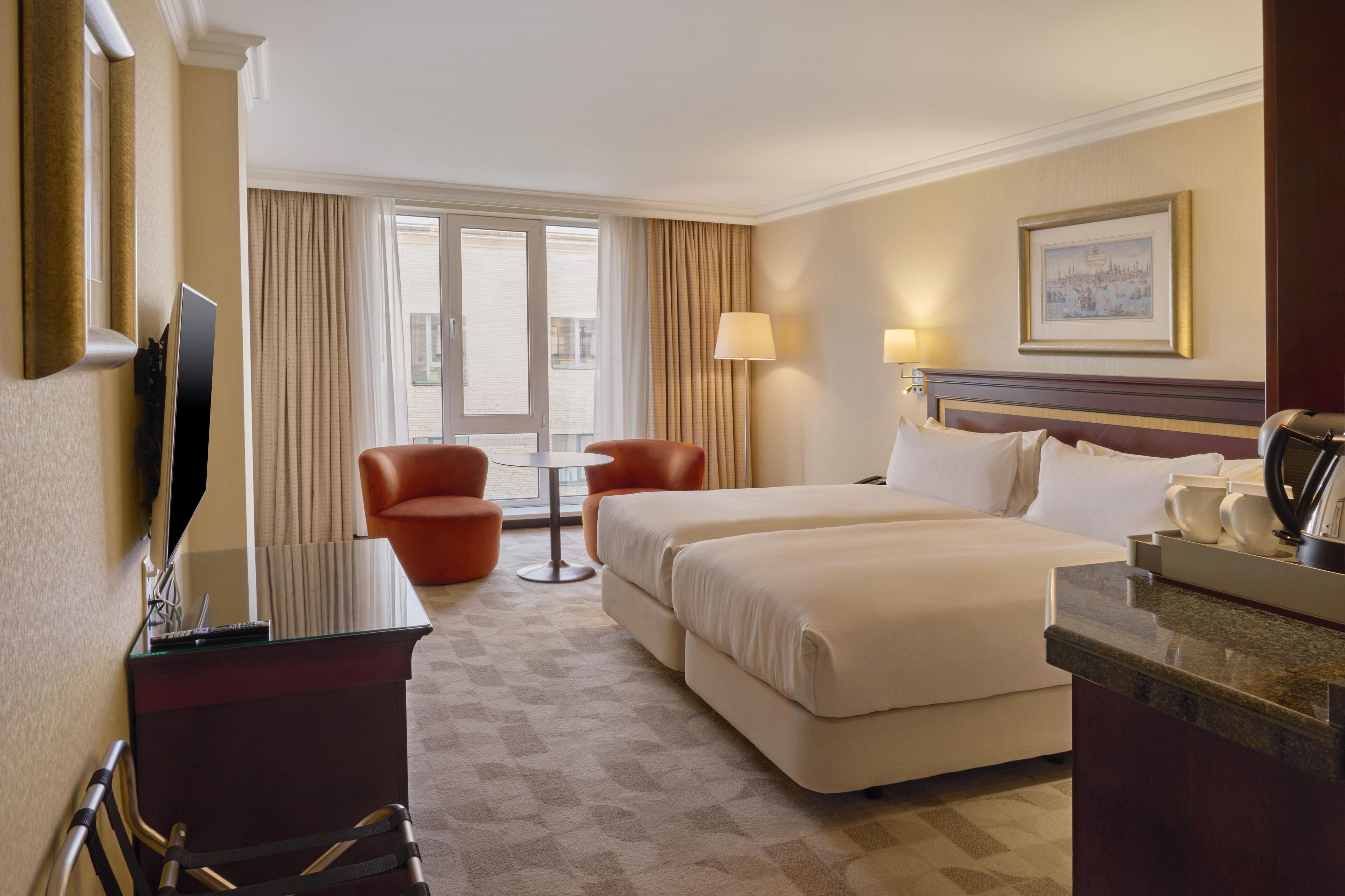 A hotel room with a large bed, a desk with a computer, a chair, a small table, and a window with curtains.