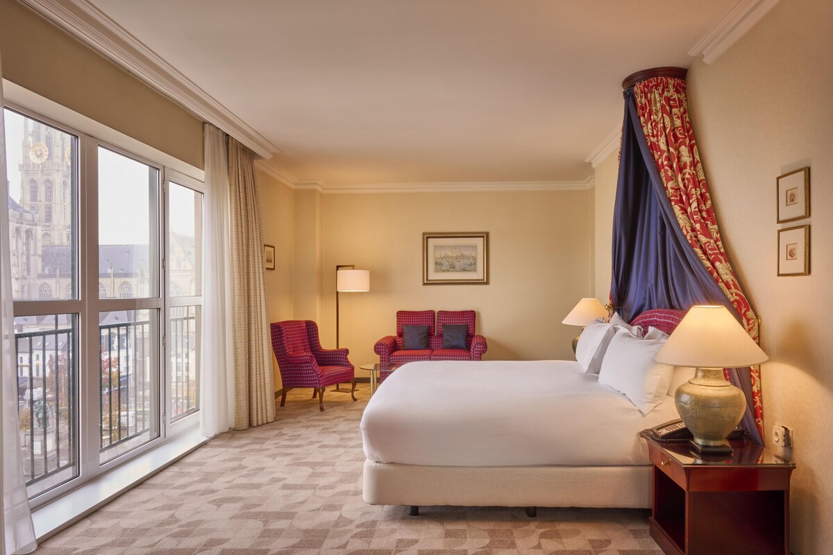 Habitación de hotel con una cama grande, dos sillones, una mesita de noche y vistas a un edificio a través de amplias ventanas.