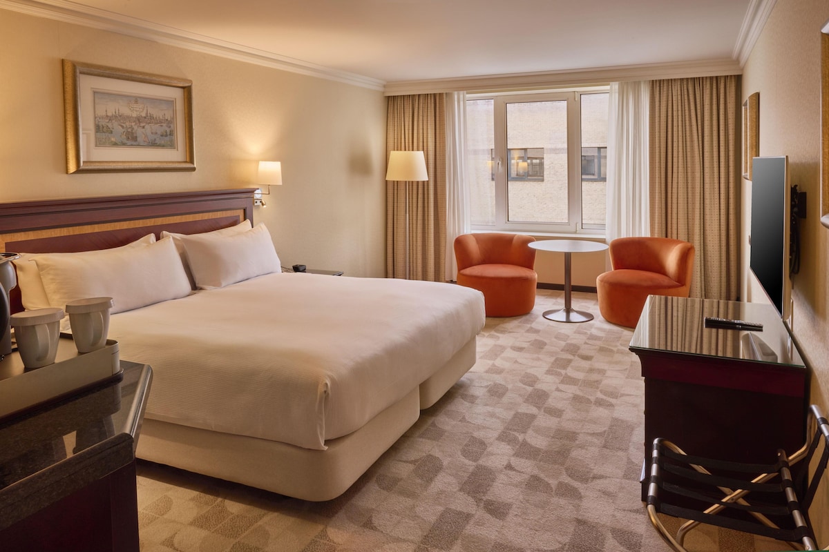 Habitación de hotel con una cama grande, dos sillones, una mesita, un escritorio con una silla, televisión y ventana con cortinas.