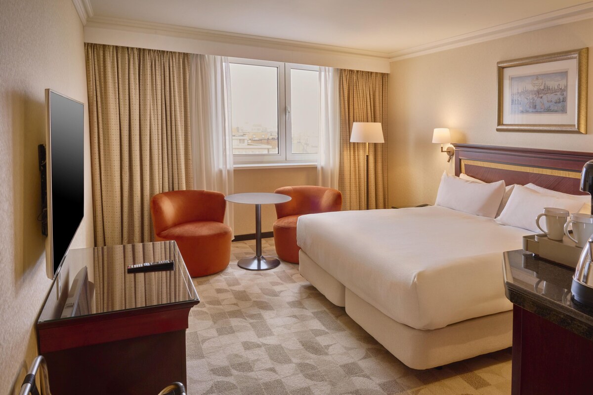 Habitación de hotel con una cama grande, dos sillones, una mesita, un televisor y una ventana con cortinas.