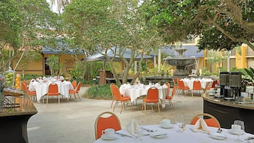 An outdoor dining area with multiple tables and chairs, surrounded by greenery and a fountain.