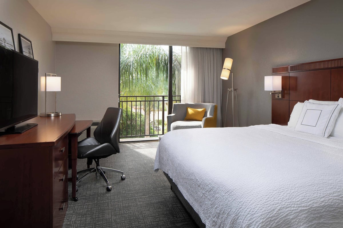 Habitación de hotel con cama, escritorio con computadora, silla, televisor y balcón con vista a follaje.