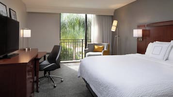 A hotel room with a bed, a desk with a computer, a chair, a TV, and a balcony with a view of greenery.