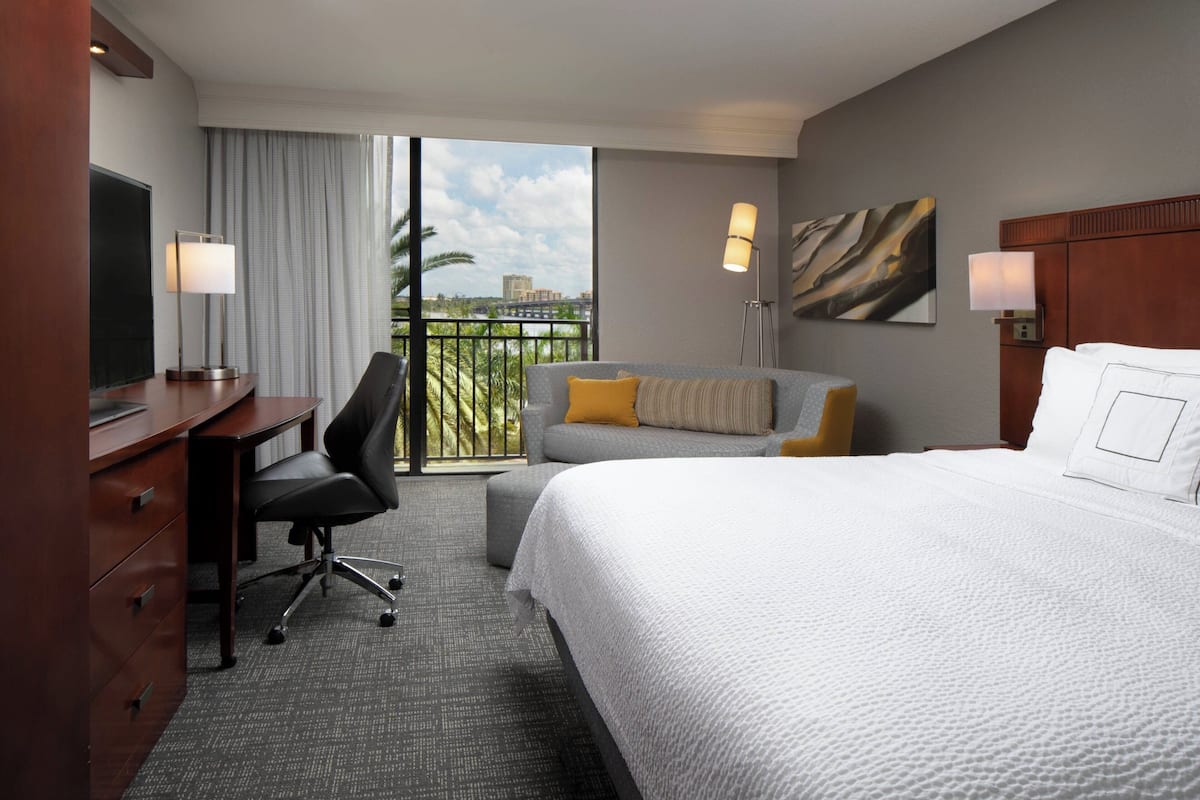 Habitación de hotel con cama, escritorio, silla, sofá, balcón con vistas y un cuadro en la pared.